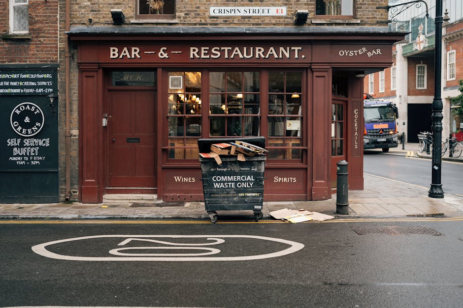 A bustling outdoor market in Notting Hill with white canopy tents and wooden stalls, surrounded by a crowd of people walking along a narrow street. The buildings are white with black and red awnings, 