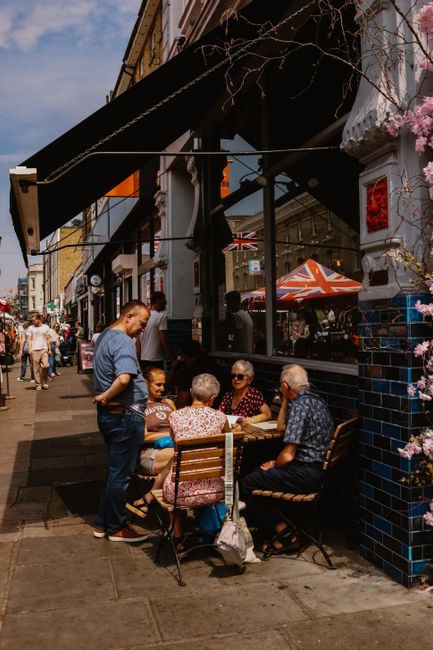 A group of five elderly women and one male waiter are seated around a small outdoor wooden table outside a cafe on a busy street in Notting Hill, London. The women are engaged in conversation, with so