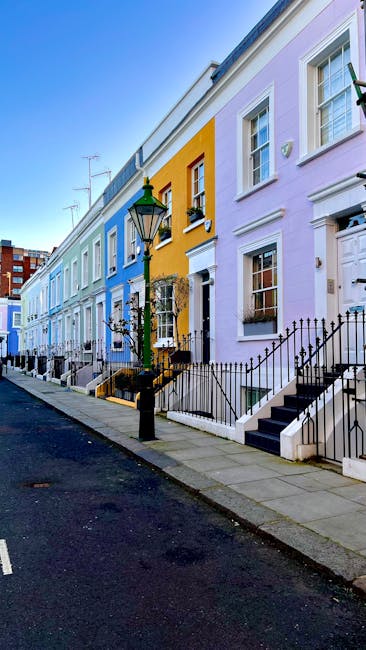 Image of a row of colorful residential townhouses on a quiet street in Notting Hill, London, painted in pastel shades of blue, yellow, and lavender. The houses feature white-framed sash windows, black cast-iron railings along the small front steps, and traditional black street lamps. The street has a clean asphalt surface with a concrete sidewalk, and the scene is illuminated by bright daylight, highlighting the vibrant exterior finishes. This setting exemplifies a well-maintained, visually appealing neighborhood that benefits from professional domestic cleaning services such as those offered by Notting Hill Cleaners, ensuring surfaces like front steps and windows are spotless and well-presented.