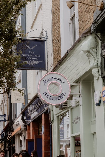 A street scene featuring various shopfronts with signage, including a prominent round neon sign shaped like a doughnut that reads 'Always a good FOR DOUGHNUTS,' and a rectangular sign for 'The Notting Hill Bookshop' hanging above storefronts. The buildings are constructed from brick and painted wood, with large windows displaying merchandise. The scene is well-lit with natural daylight, and the area appears lively with pedestrians visible at the bottom of the image. This image captures the exterior of a commercial street in Notting Hill, with shops that enhance the area's vibrant atmosphere relevant to domestic and commercial cleaning contexts emphasized by Notting Hill Cleaners.