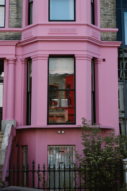 Image of a row of colorful residential townhouses on a quiet street in Notting Hill, London, painted in pastel shades of blue, yellow, and lavender. The houses feature white-framed sash windows, black cast-iron railings along the small front steps, and traditional black street lamps. The street has a clean asphalt surface with a concrete sidewalk, and the scene is illuminated by bright daylight, highlighting the vibrant exterior finishes. This setting exemplifies a well-maintained, visually appealing neighborhood that benefits from professional domestic cleaning services such as those offered by Notting Hill Cleaners, ensuring surfaces like front steps and windows are spotless and well-presented.