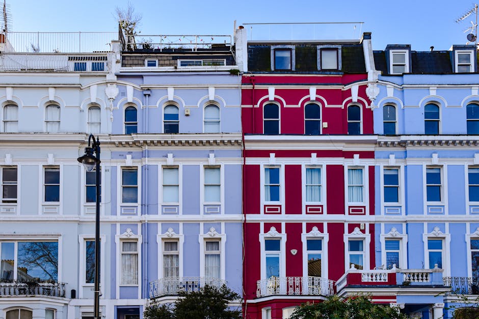 A row of Victorian-style residential buildings with colorful facades in pastel shades of pink, blue, peach, and white, featuring large bay windows, decorative moldings, and small balconies with black