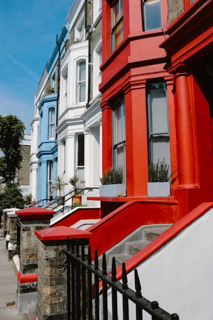 A row of Victorian-style terraced houses in Notting Hill with colorful facades including shades of pink, white, blue, and teal. The buildings feature large sash windows, some with flower boxes or pott